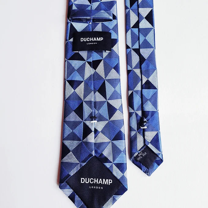 Back view of the Duchamp London Blue Geometric Silk Tie showing both Duchamp labels and the inner lining of the wide blade tip. The intricate geometric design and craftsmanship are evident in the premium silk fabric.